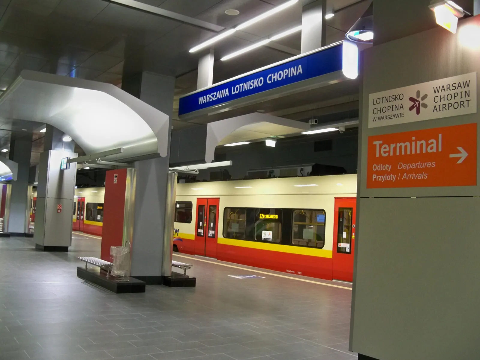 The underground railway platform at Warsaw Chopin Airport, a red and yellow commuter train at the edge and an orange sign overhead pointing the way up to the terminal