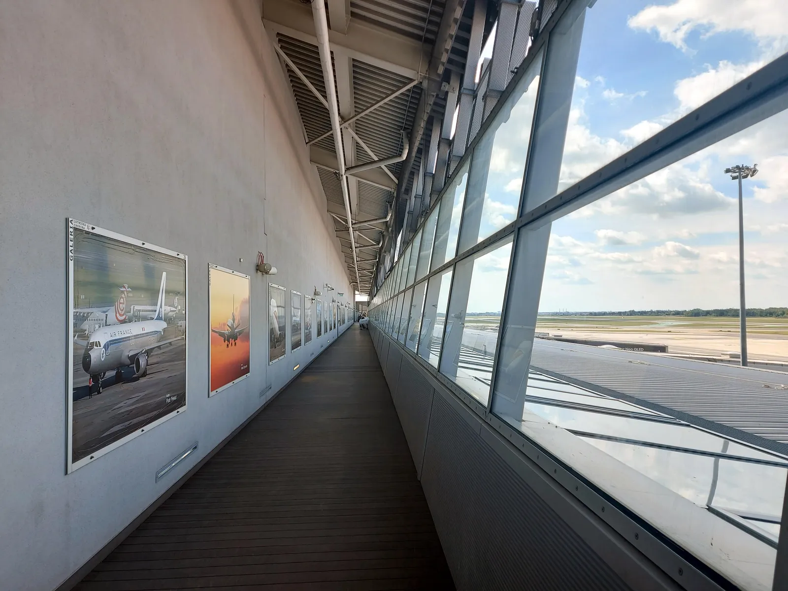 The observation deck at Warsaw Chopin Airport, a long narrow gallery glazed floor to ceiling down one side looking out over the apron and taxiways, with framed aviation photographs hung along the opposite wall