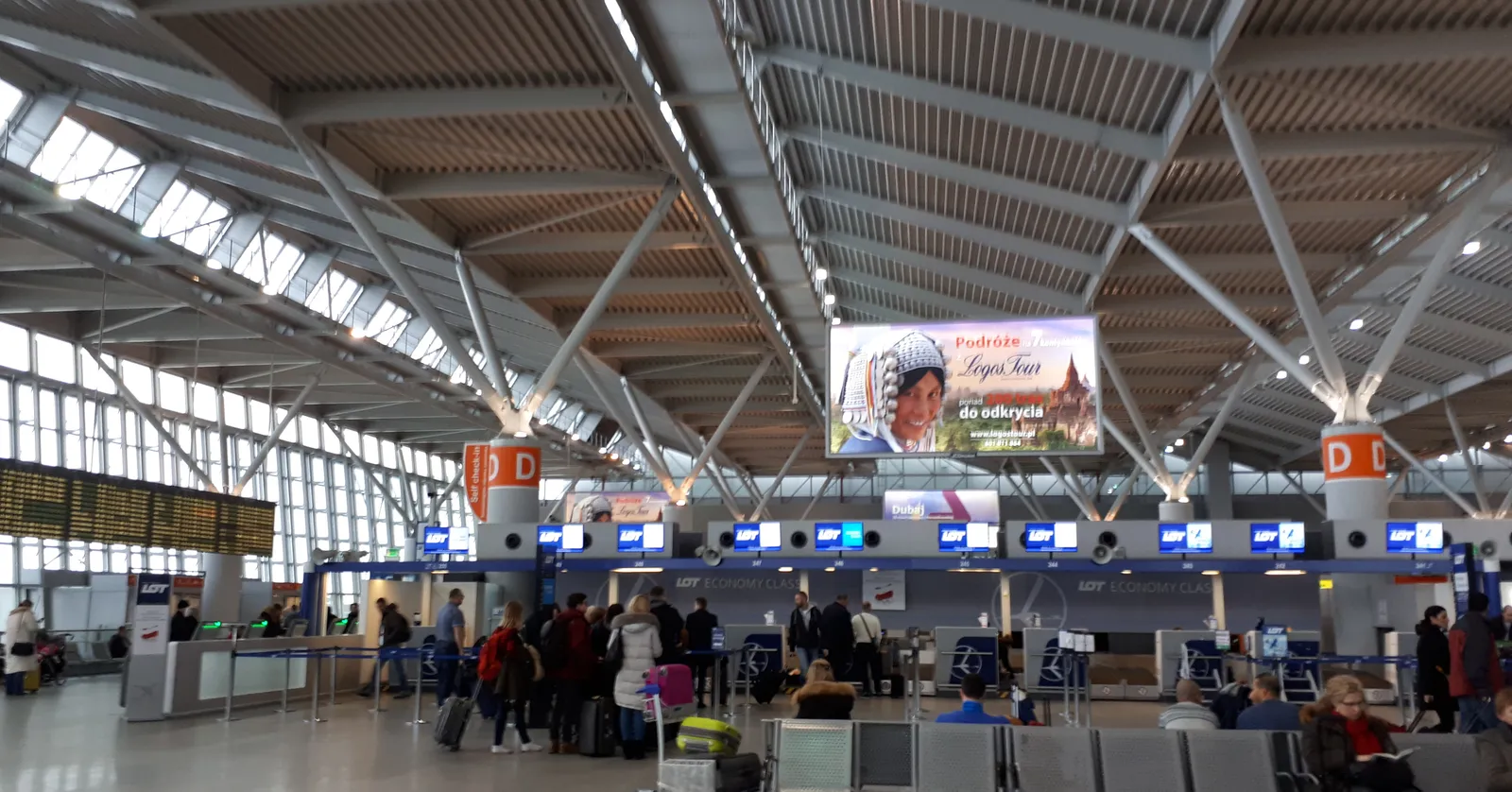 The departures hall at Warsaw Chopin Airport, a long steel-trussed roof over rows of check-in desks lettered D, with passengers and trolleys queuing beneath a banner carrying a portrait of Chopin