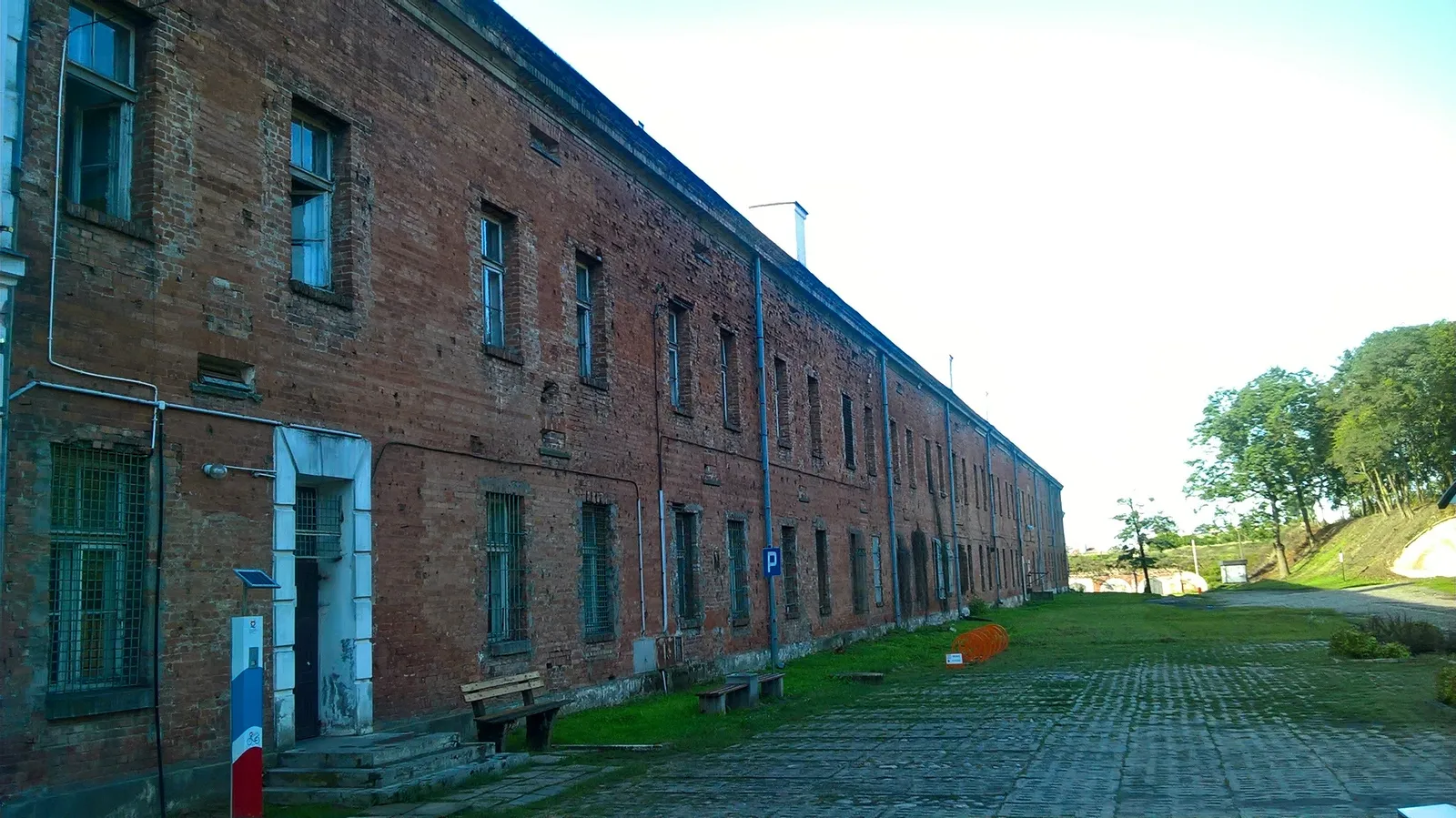 A long two-storey red brick barracks range at Modlin Fortress running away into the distance in an unbroken line, grass and a cobbled track alongside it