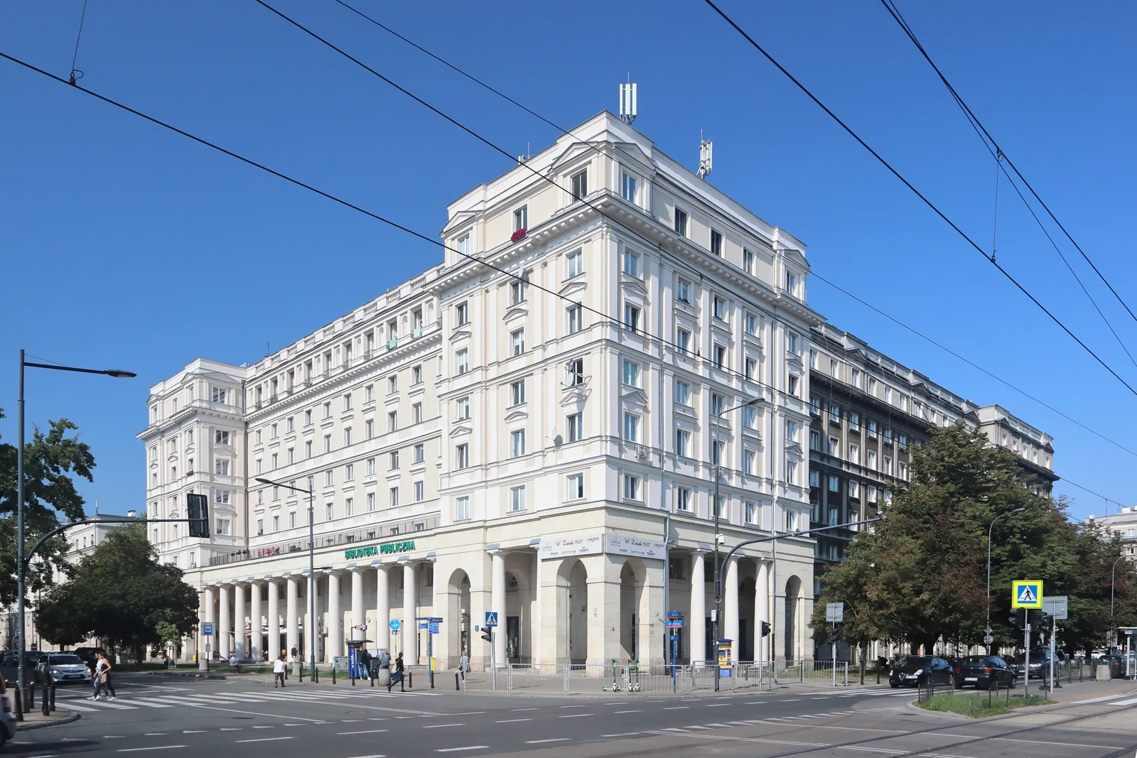 A large white 1950s apartment block on Anielewicza in Warsaw's Muranów district, its corner carried on a colonnade, tram wires strung across the junction in front of it