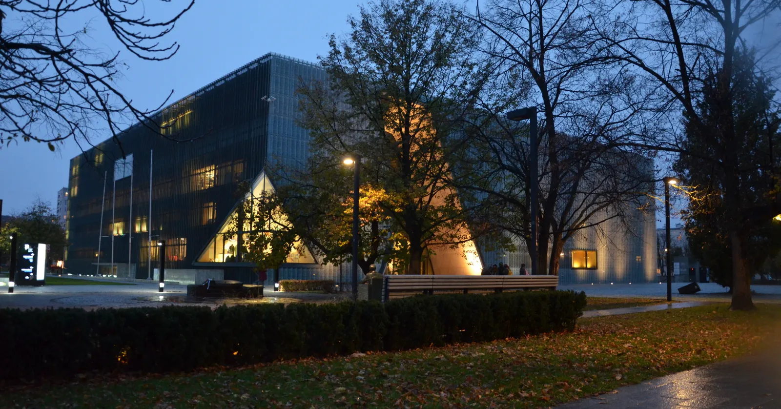 The POLIN museum in Warsaw at dusk, a low glass and copper box lit from within, with the tall curved slit of its entrance cut through the middle of the facade