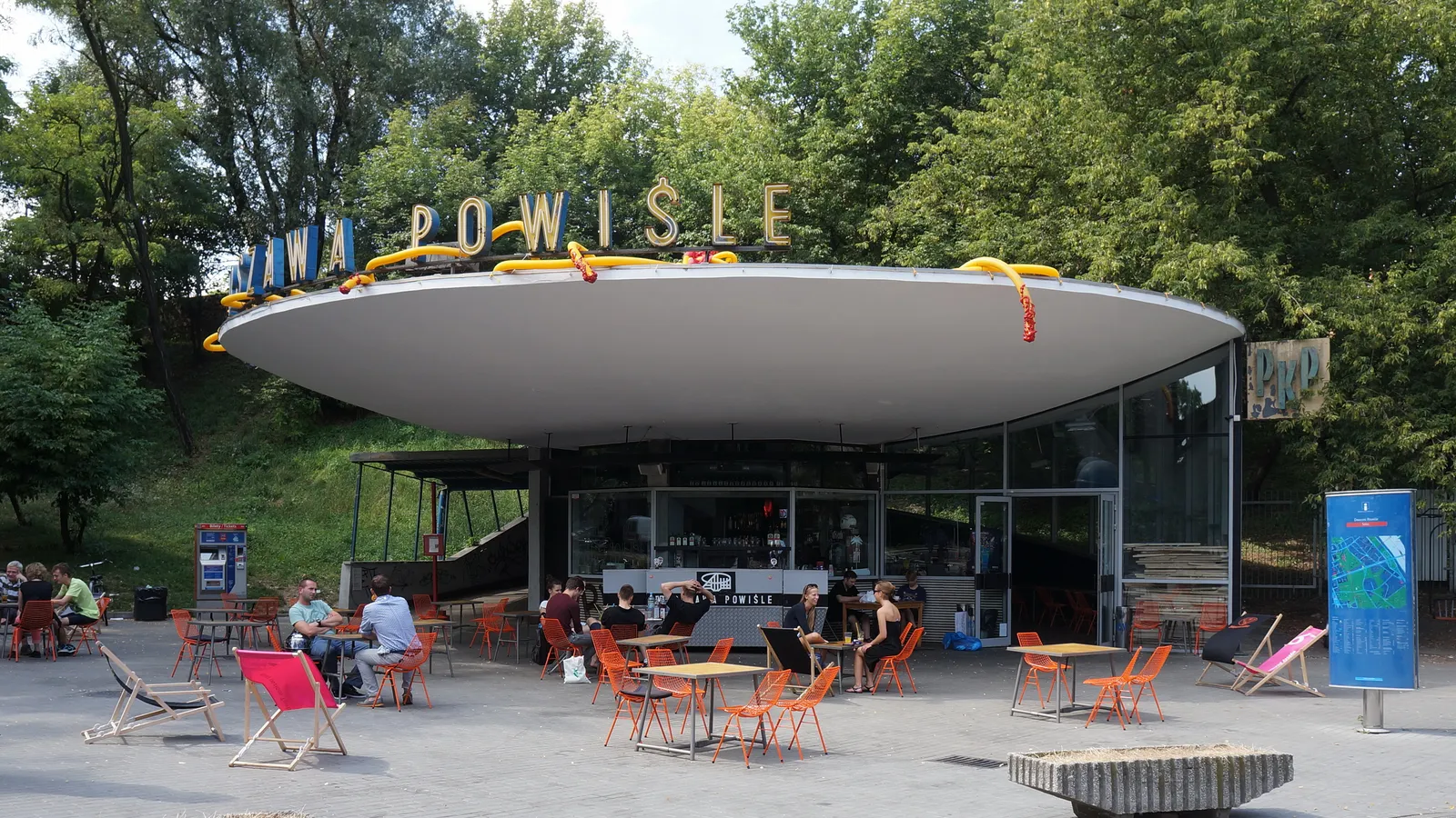 The old ticket pavilion at Warszawa Powiśle station, a small glazed drum under a wide saucer-shaped concrete roof with the station name in metal letters along its rim, café tables and deckchairs set out in front