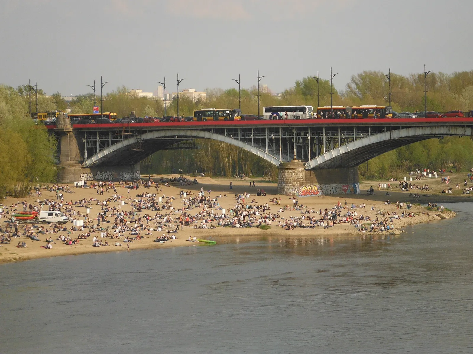 The Vistula in Warsaw seen from the water, the steel arches of the Poniatowski Bridge crossing it and a wide sandy beach crowded with people on the undeveloped far bank
