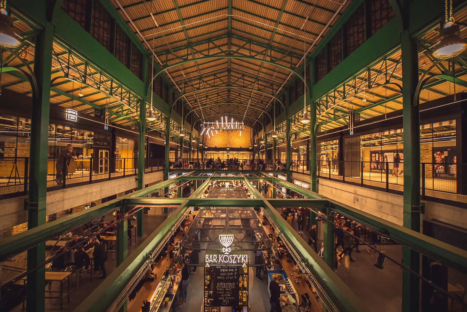 Inside Hala Koszyki in Warsaw, a green-painted iron frame carrying a pitched glass roof over a two-level hall with food counters and long shared tables on the floor below