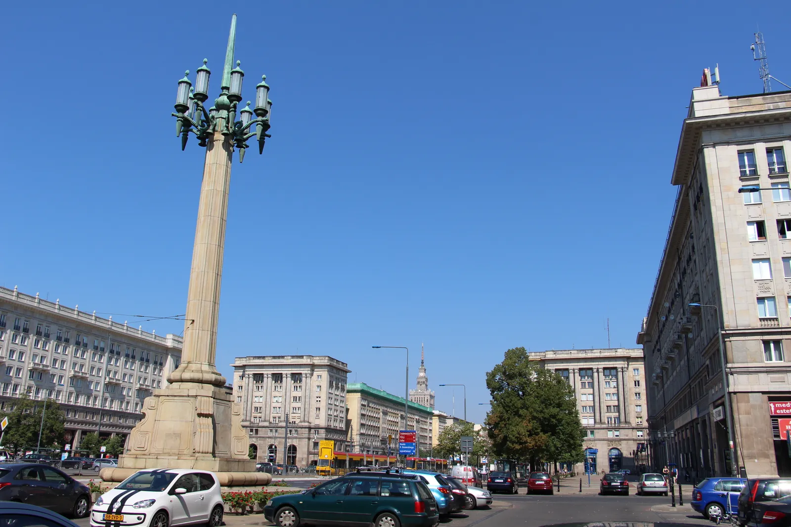 Plac Konstytucji in Warsaw, a tall stone candelabrum carrying a cluster of lanterns in the foreground and the heavy 1950s apartment blocks of the Marszałkowska district ranged around the square behind it