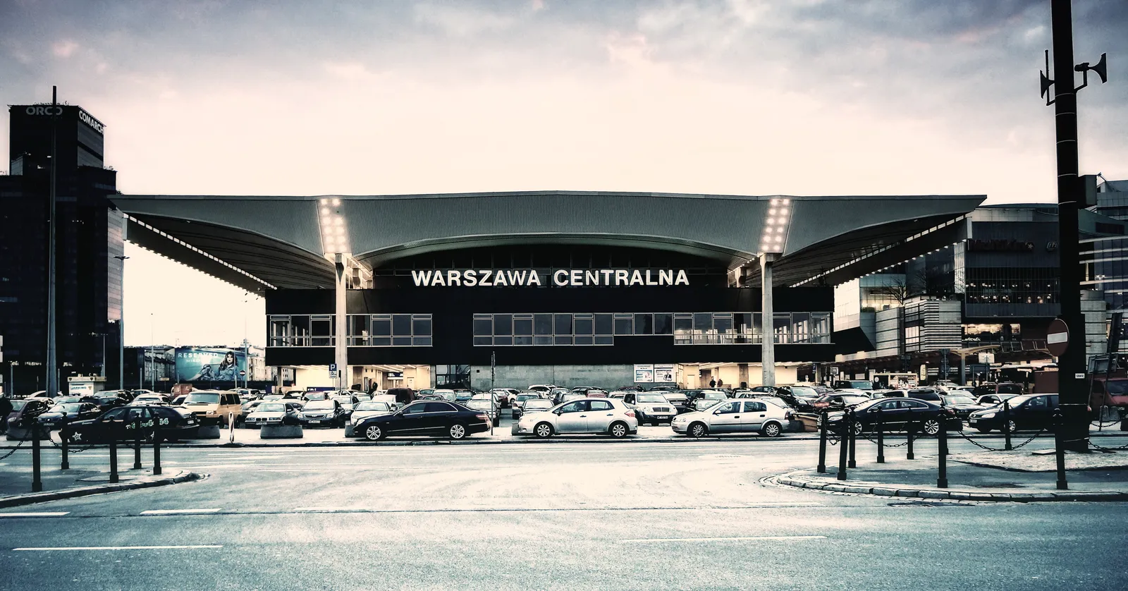 The front of Warszawa Centralna station, a low 1970s block under a wide cantilevered concrete canopy with the name lettered across it, cars parked along the forecourt