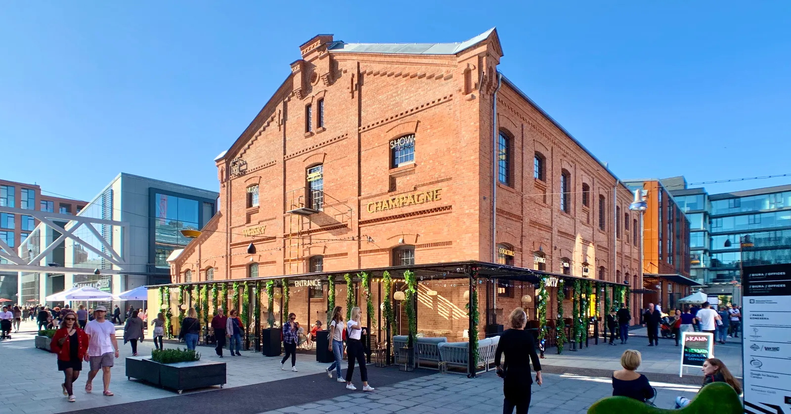 The restored Koneser distillery in Warsaw's Praga district, a red brick industrial building with CHAMPAGNE lettered across its gable, opening onto a paved public square with people crossing it