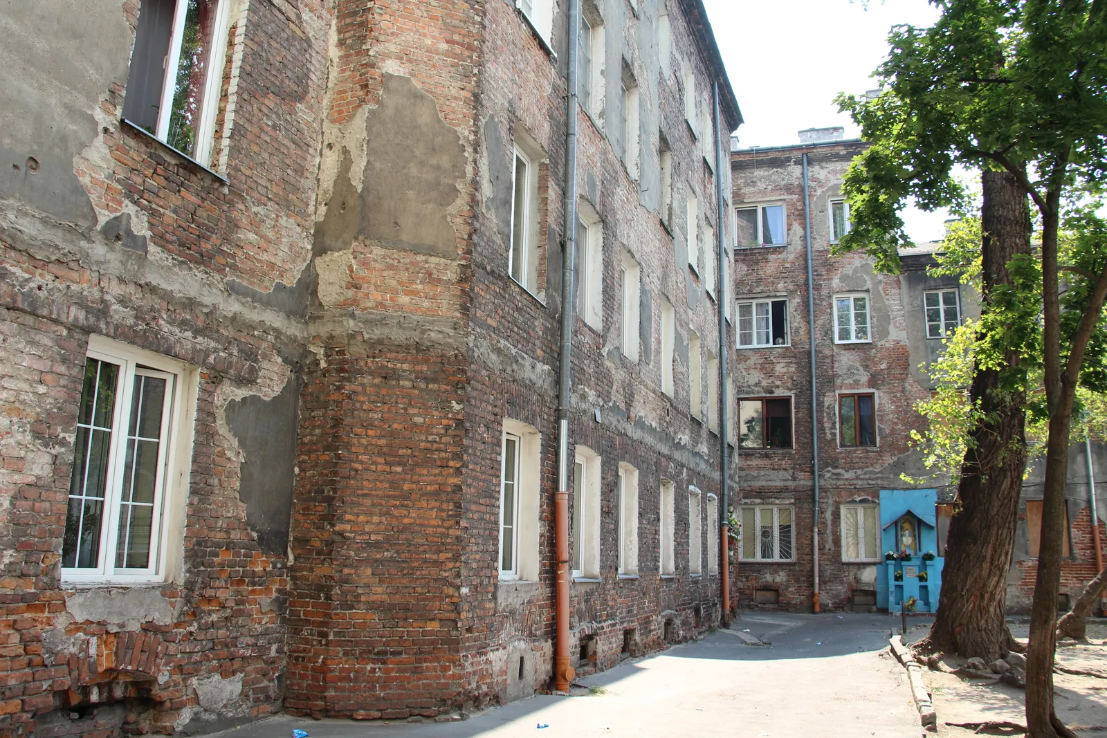 A courtyard off Ząbkowska Street in Warsaw's Praga district, tenement walls with plaster falling away to bare brick, and a small blue shrine set against the far wall under a tree