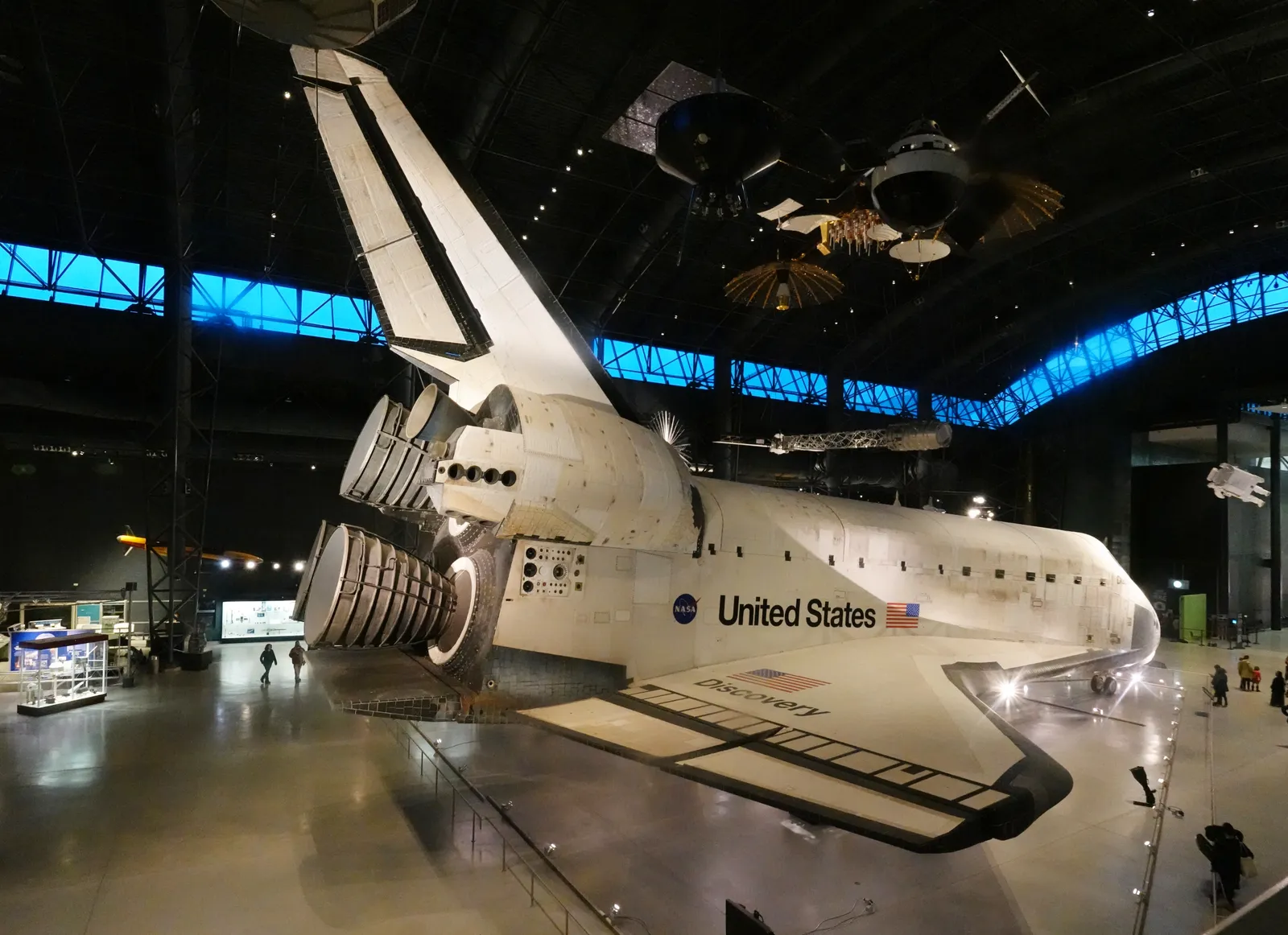 Space Shuttle Discovery on display at the Udvar-Hazy Center, its scorched heat-shield tiles and open engine bells filling a hangar with satellites hanging above it