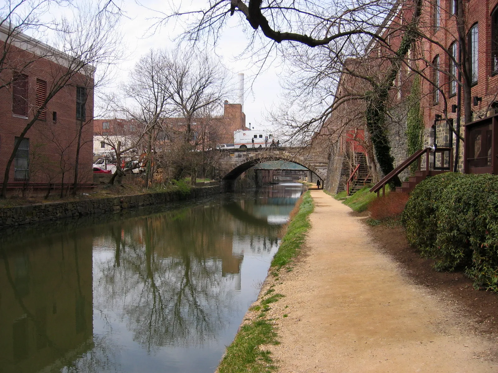 The Chesapeake and Ohio Canal running through Georgetown between brick warehouses, with a gravel towpath along one bank and a stone arch bridge ahead