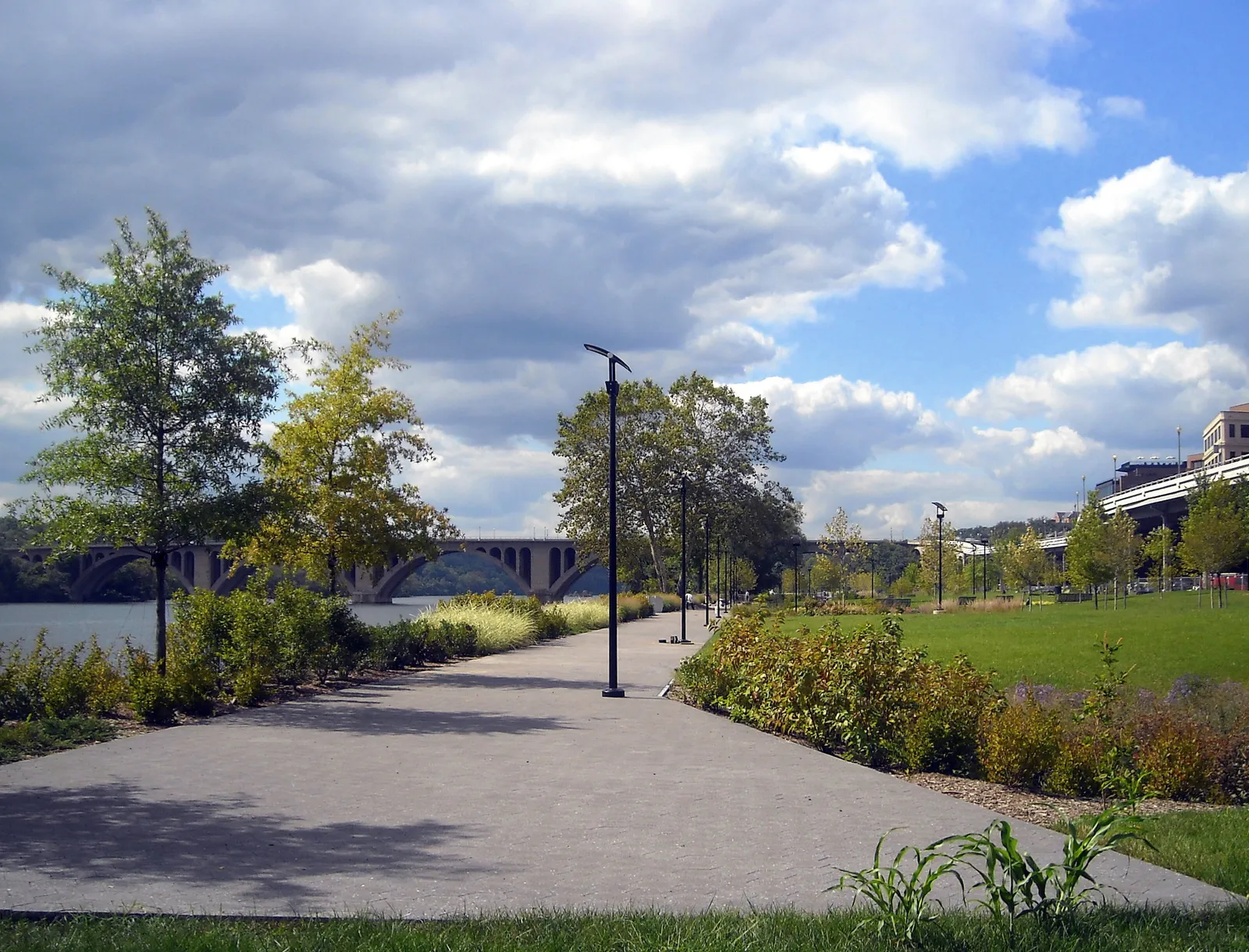 The paved riverside path through Georgetown Waterfront Park in Washington, with young trees, lawn and the arches of Key Bridge in the distance