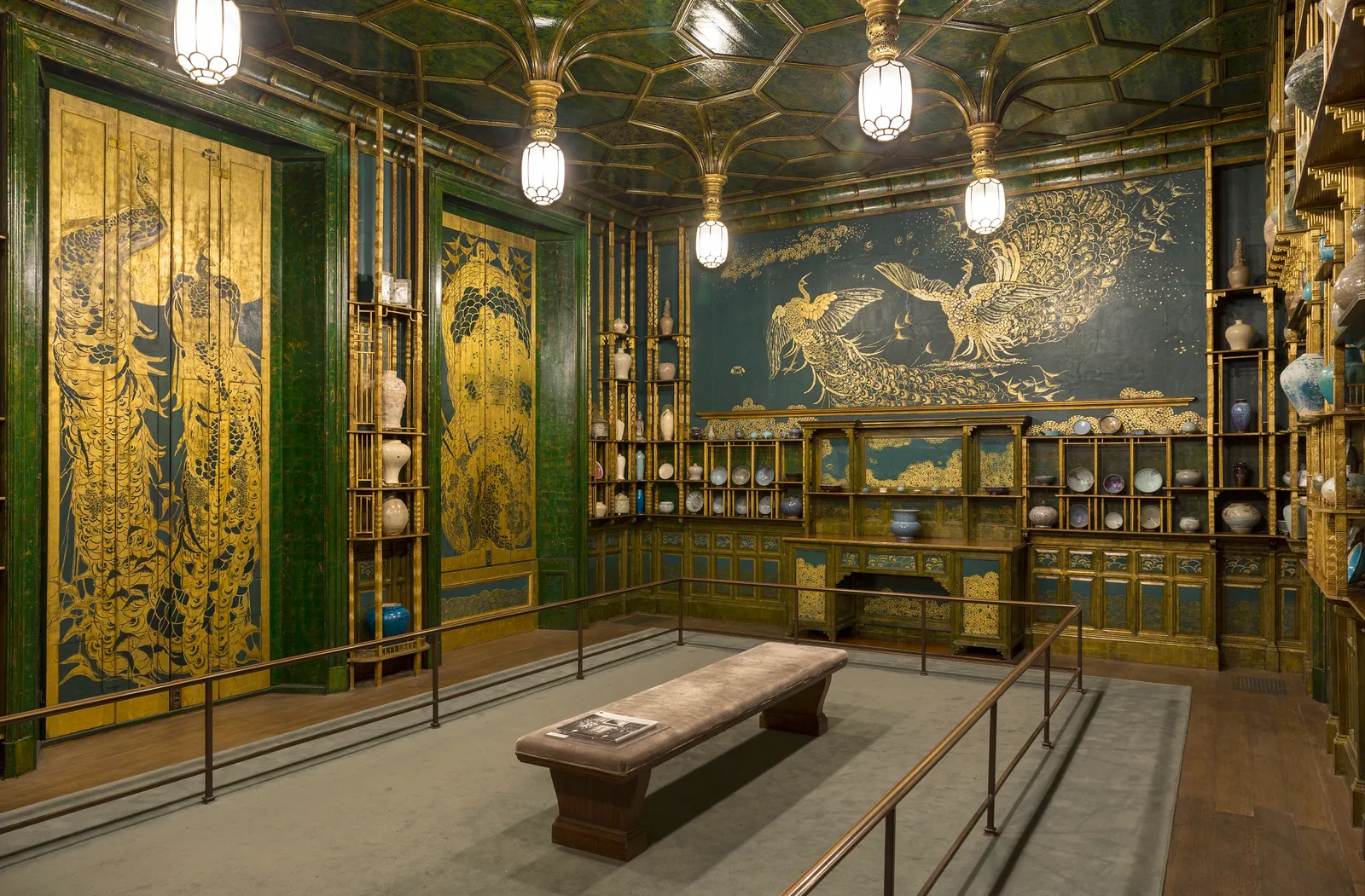 The Peacock Room at the Freer Gallery in Washington, its walls and shutters painted with gold peacocks over blue-green, lined with shelving for porcelain and lit by hanging lamps