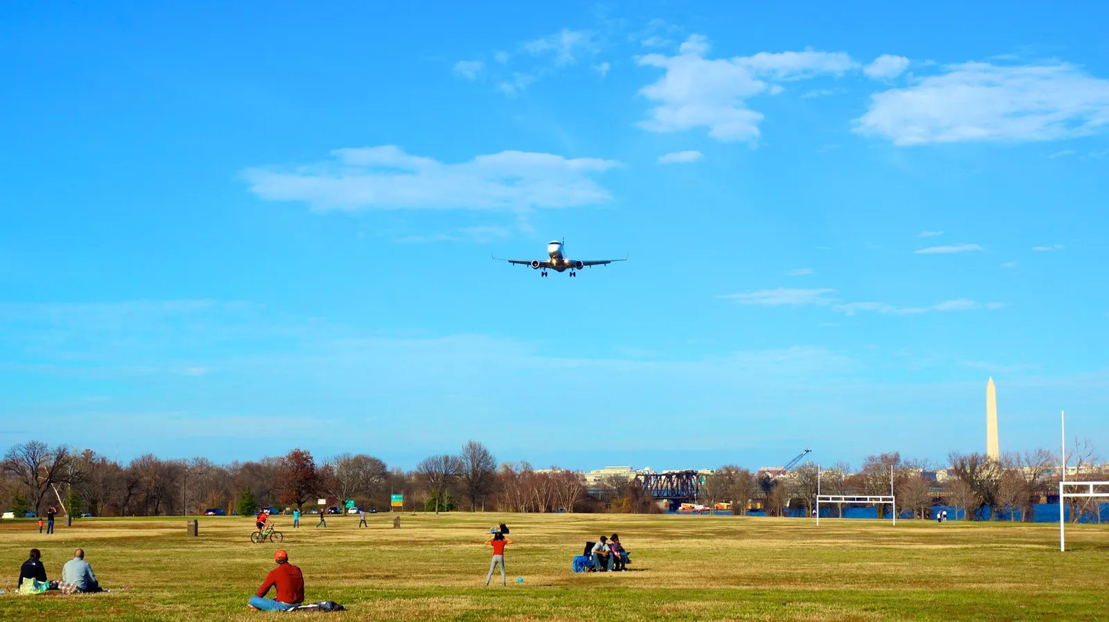 An airliner on final approach low over the open lawn at Gravelly Point in Arlington, with people sitting on the grass below and the Washington Monument on the skyline behind