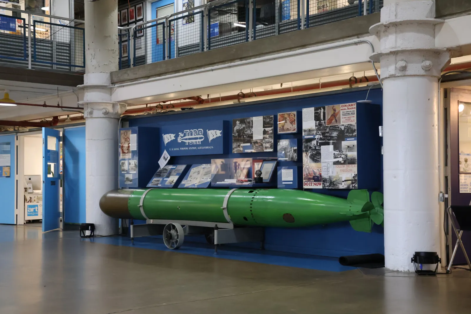 A green torpedo on a wheeled cradle displayed inside the Torpedo Factory Art Center in Alexandria, with white cast-iron columns and studio balconies above it