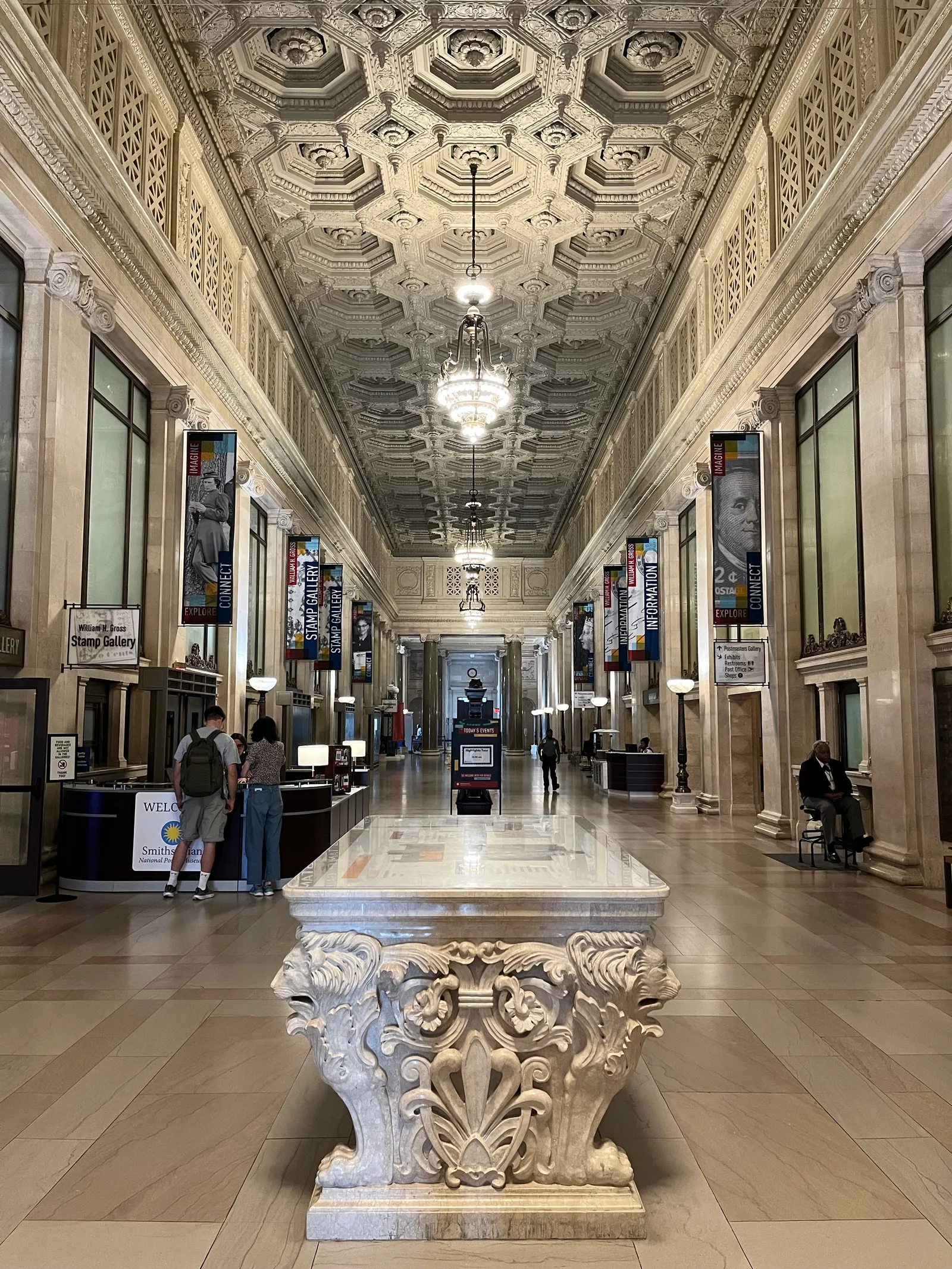 The lobby of the National Postal Museum in Washington, a long marble hall under a deeply coffered plaster ceiling with chandeliers and exhibition banners along both sides