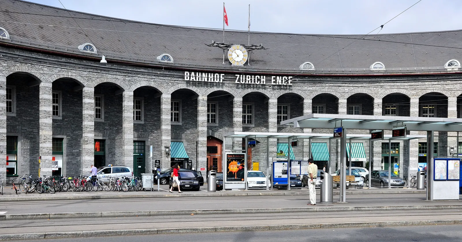 The curved granite front of Zürich Enge station on Tessinerplatz, a long arcaded facade of round-headed openings under a low grey roof with a large clock above the central entrance