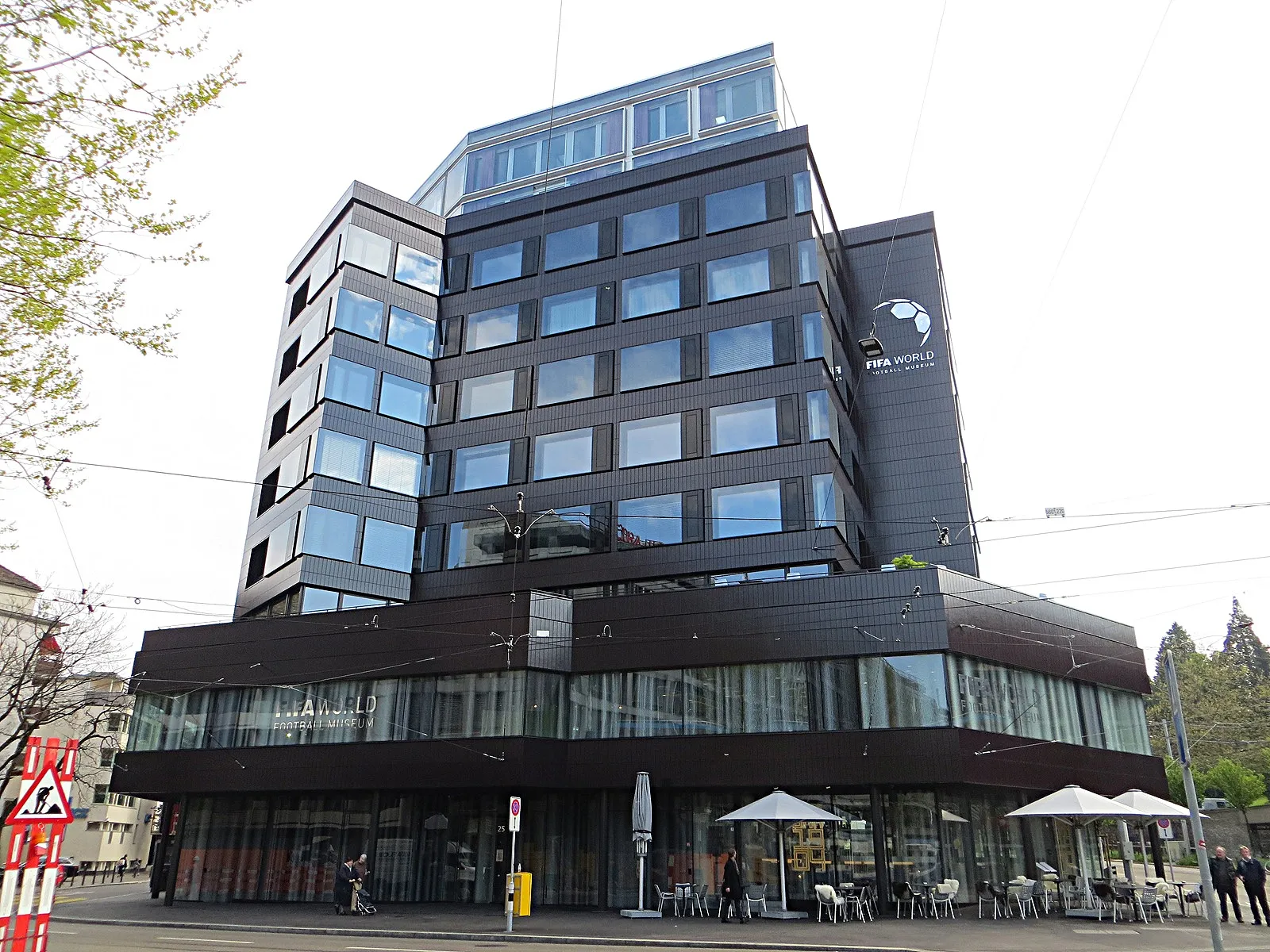 The dark-clad corner building housing the FIFA Museum in Zurich, with ribbon windows on the upper floors, a projecting glazed ground floor and cafe parasols on the pavement outside