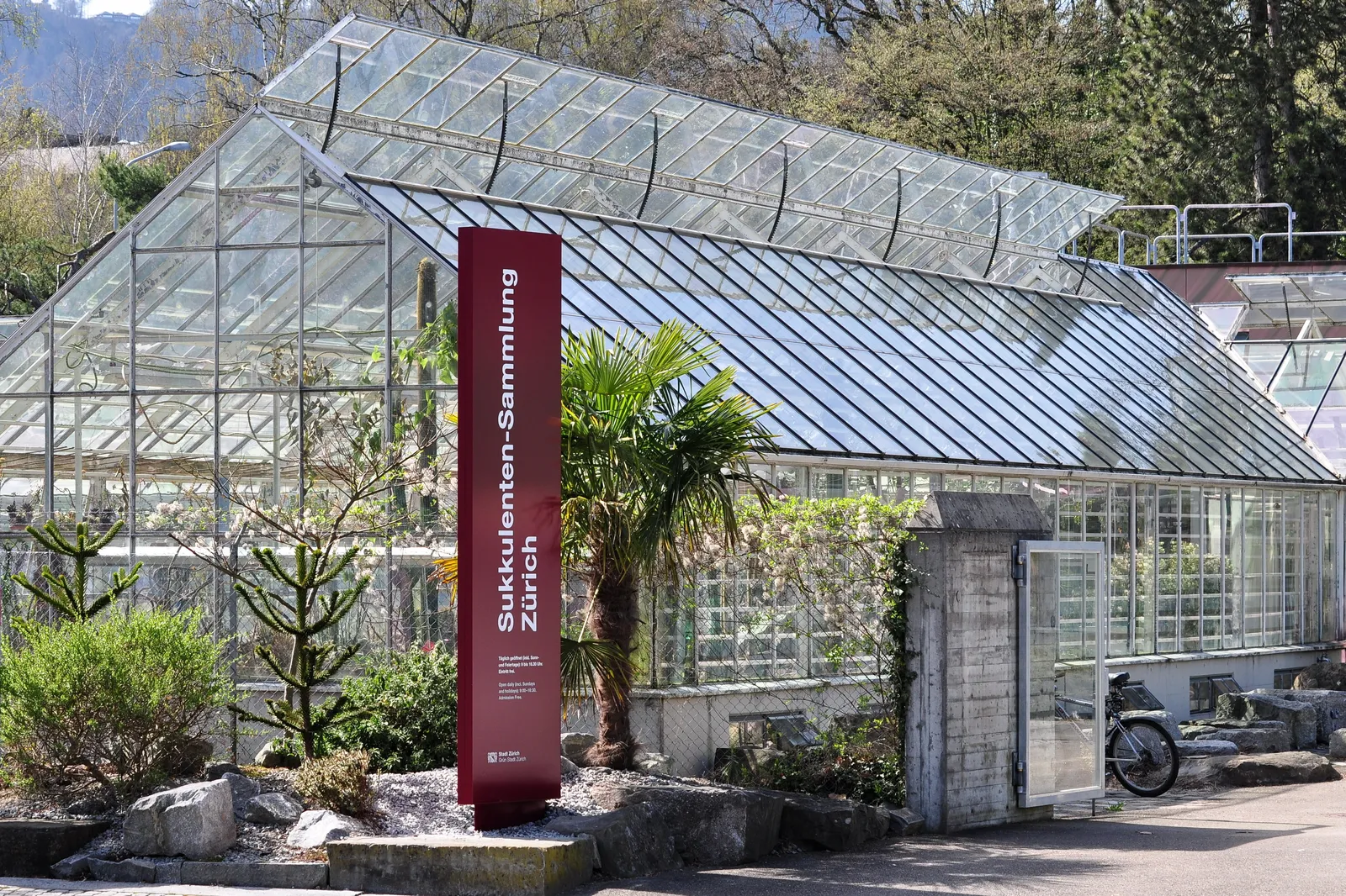 The glasshouses of Zurich's succulent plant collection on the Mythenquai, low pitched greenhouse roofs behind a rockery of cacti and palms, with the collection's name on a red sign at the entrance