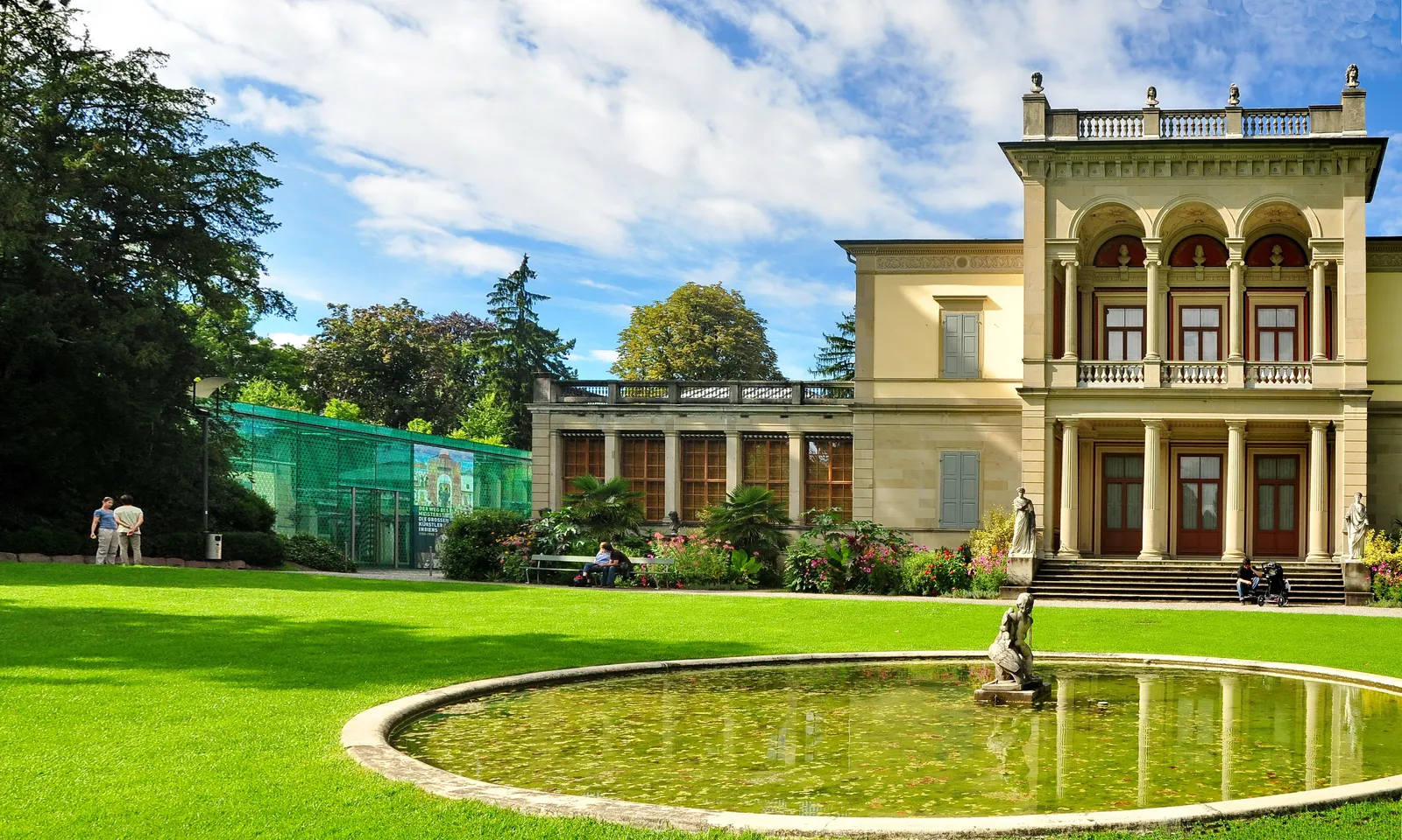 The Villa Wesendonck in Zurich's Rieterpark, a pale classical villa with a loggia of arches, seen across a lawn with a circular ornamental pond in front and the green glass entrance pavilion of the Museum Rietberg to the left