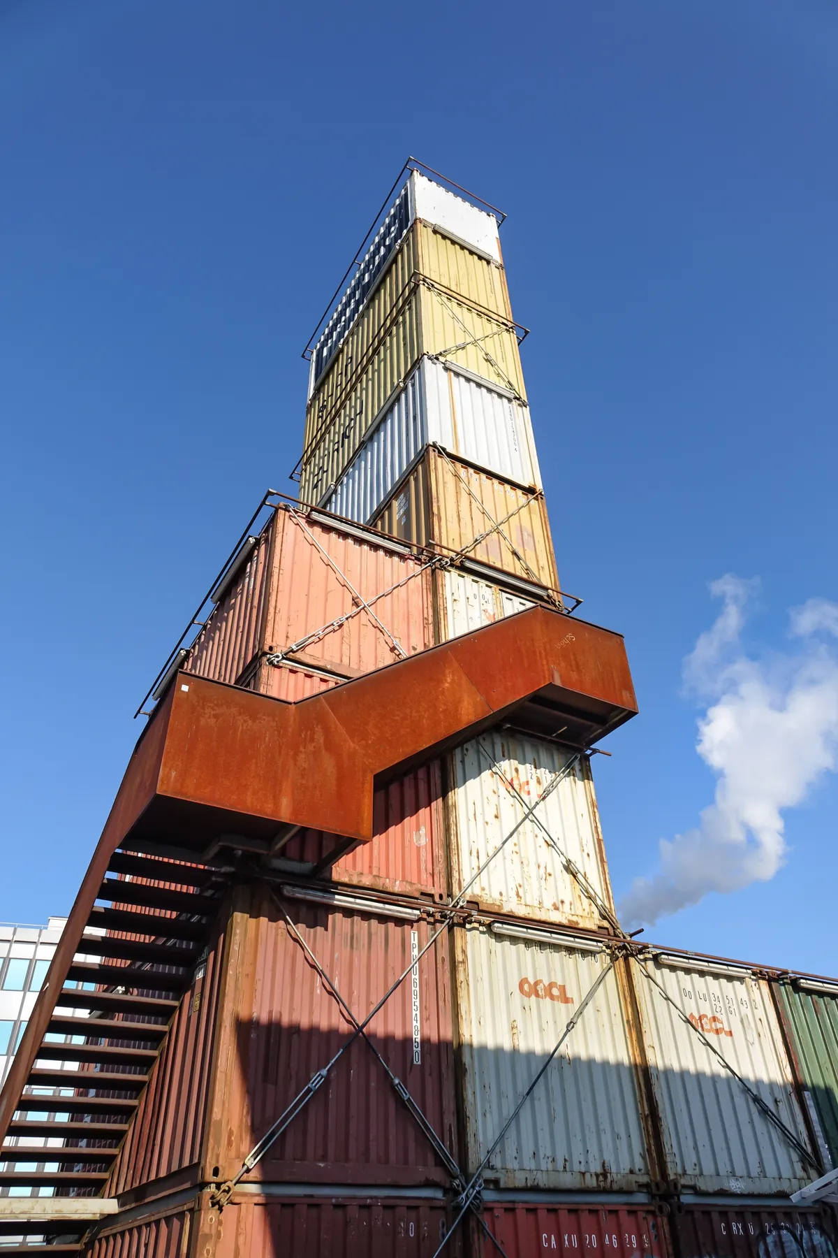 The Freitag Tower in Zurich seen from below, a narrow stack of rusted and painted shipping containers with an external steel staircase up one side and a cantilevered container jutting out partway up, against a blue sky