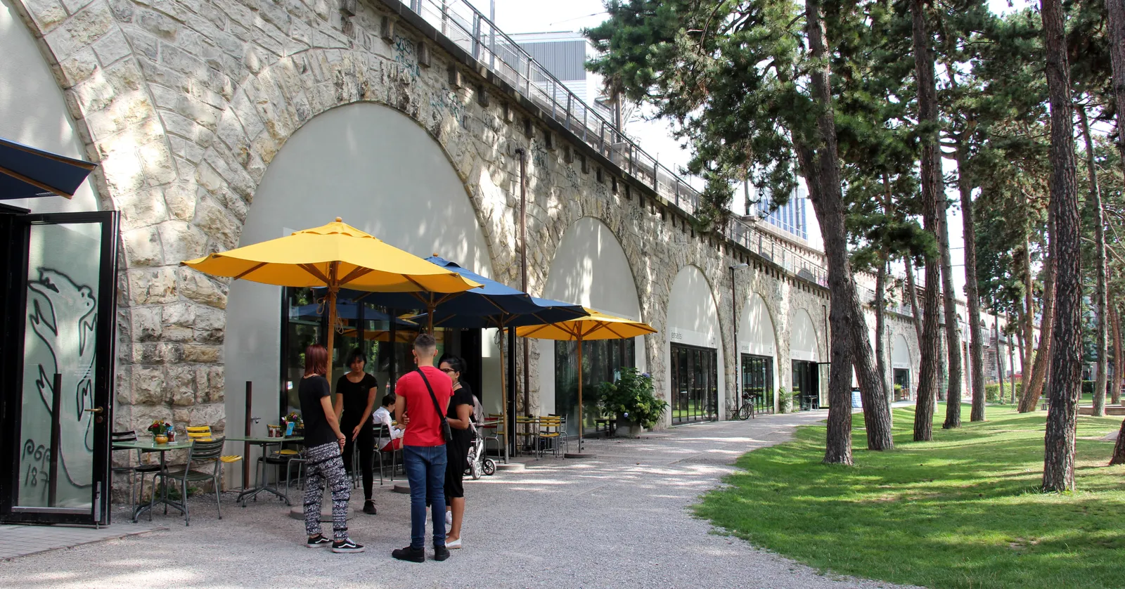 The Im Viadukt arcade in Zurich, a long line of stone railway arches filled in with shopfronts, yellow parasols and cafe tables outside one of them, and a gravel path running alongside under tall trees