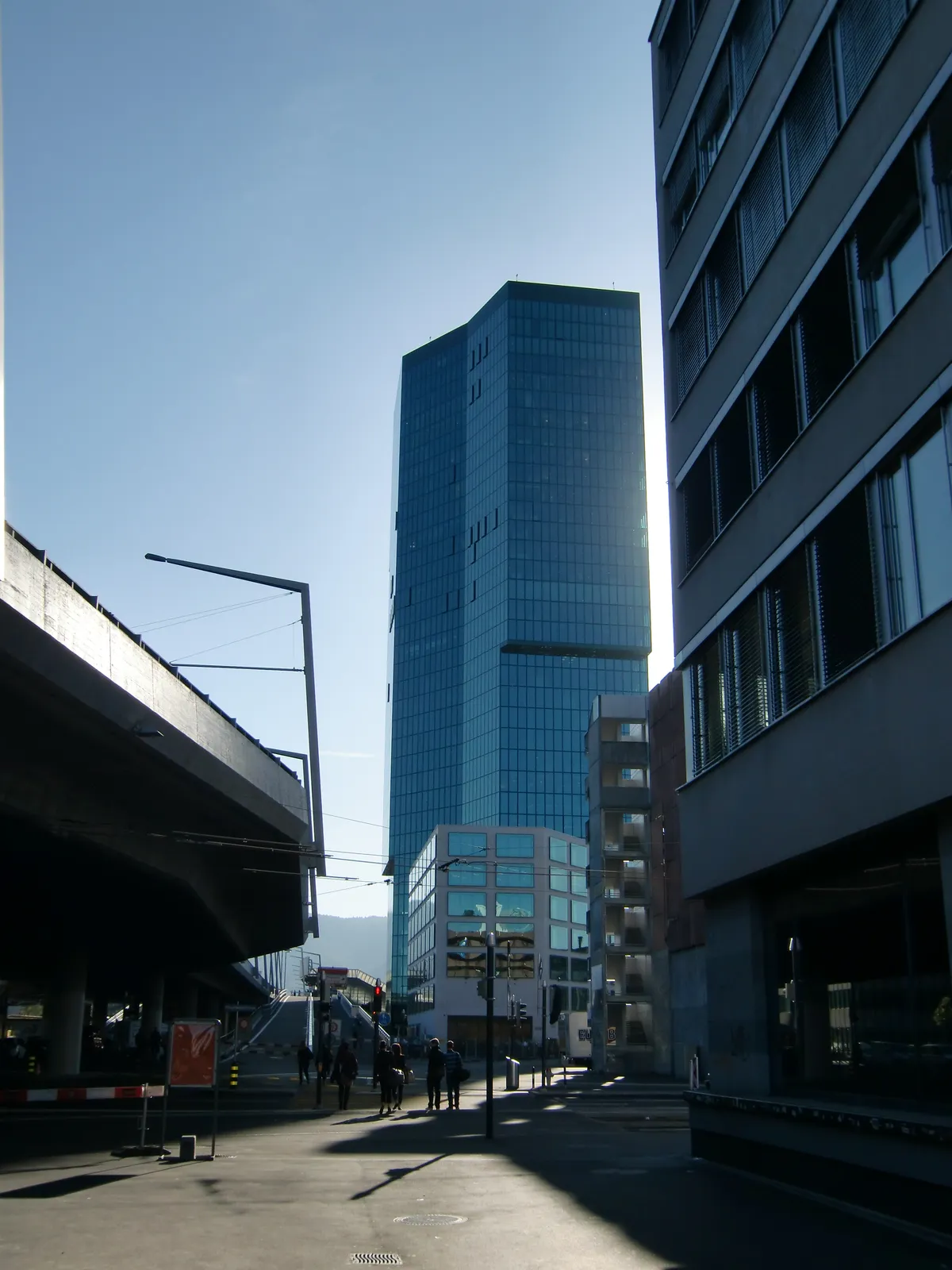 The Prime Tower in Zurich, a dark green glass office tower with an irregular polygonal plan, seen down a street with the concrete deck of the Hardbrücke road viaduct in the foreground