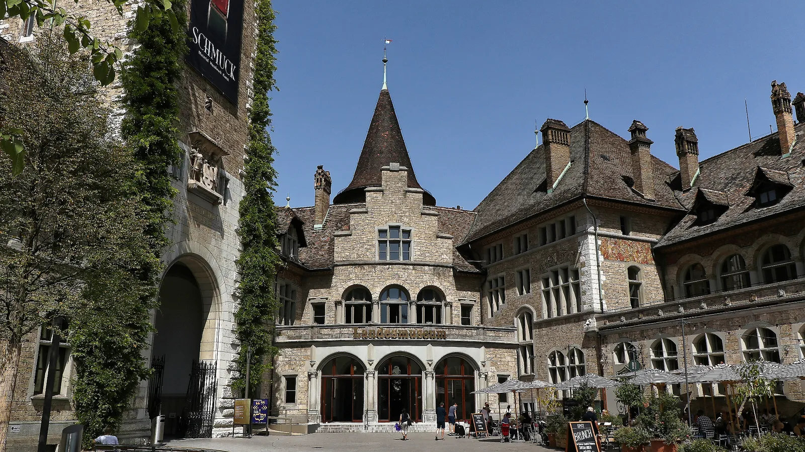 The inner courtyard of the Swiss National Museum in Zurich, a stone building with a steep pointed tower, stepped gables and an arcaded entrance, with tables and parasols set out on the gravel in front of it