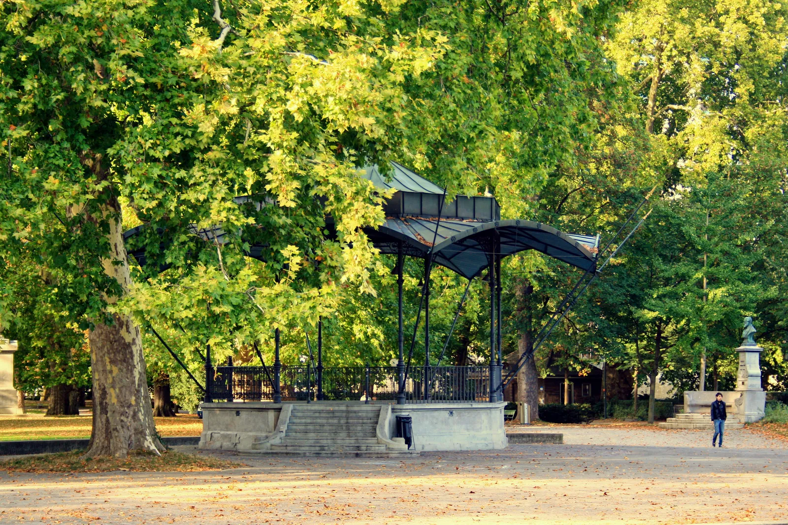 The wrought-iron music pavilion in the Platzspitz park in Zurich, standing on a low stone platform under tall plane trees, with a gravel path curving past it and a single person walking by