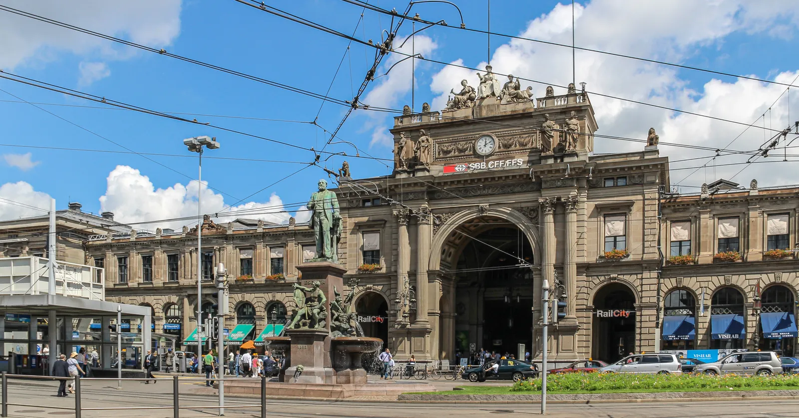 The sandstone front of Zürich Hauptbahnhof on Bahnhofplatz, a wide neo-Renaissance facade with a tall central arch under a clock, allegorical statues along the roofline, and the Alfred Escher monument and its fountain in front of it