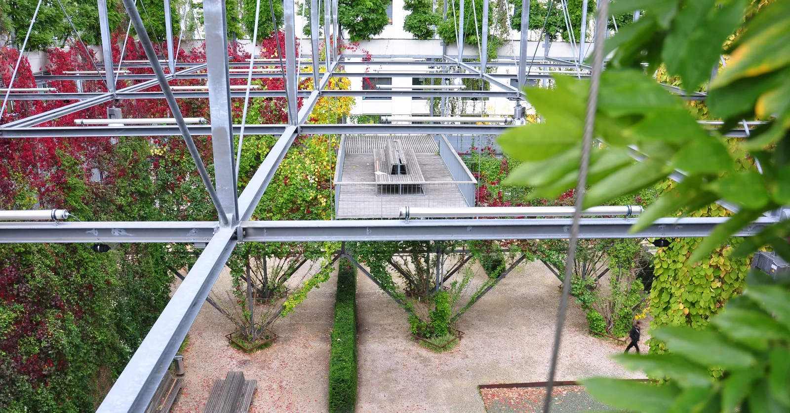 Inside the MFO-Park in Zurich Oerlikon, looking down through the levels of an open steel frame hung with green and red climbing plants, with wooden decking on an upper platform and a gravel courtyard below