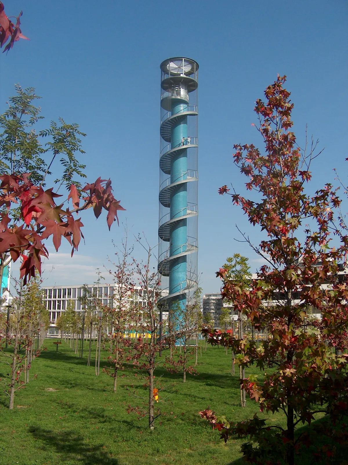 The blue concrete viewing tower in Oerliker Park in Zurich, a slim shaft wrapped in a spiral steel staircase and a fine mesh net, rising above young trees with apartment blocks behind