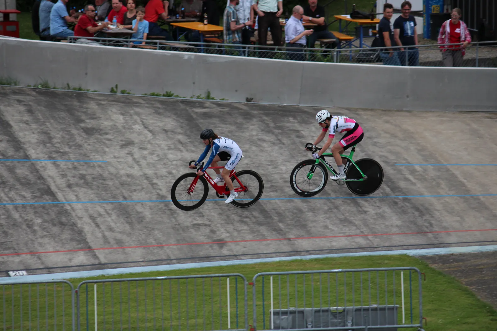 Two track cyclists riding high on the steeply banked concrete curve of the open-air velodrome at Zurich Oerlikon, with spectators at tables above the rail behind them