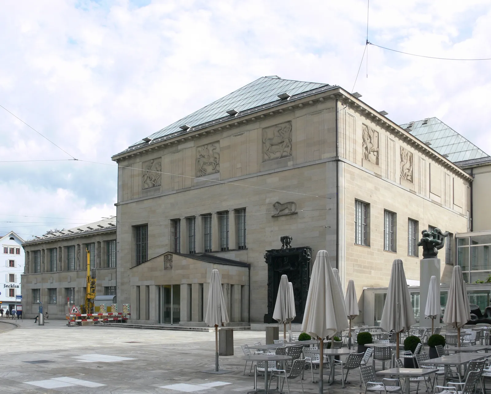 The original Moser building of the Kunsthaus Zürich on Heimplatz, a squared sandstone block with a green copper roof and carved relief panels along the upper wall, with cafe tables and closed parasols on the square in front