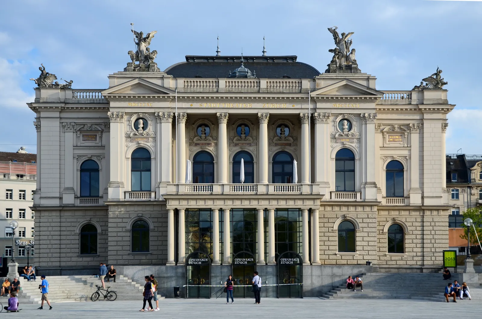 The Zurich Opera House seen across Sechseläutenplatz, a symmetrical stone facade with a columned portico, arched windows and winged statues along the roofline, with people crossing the paved square in front of it