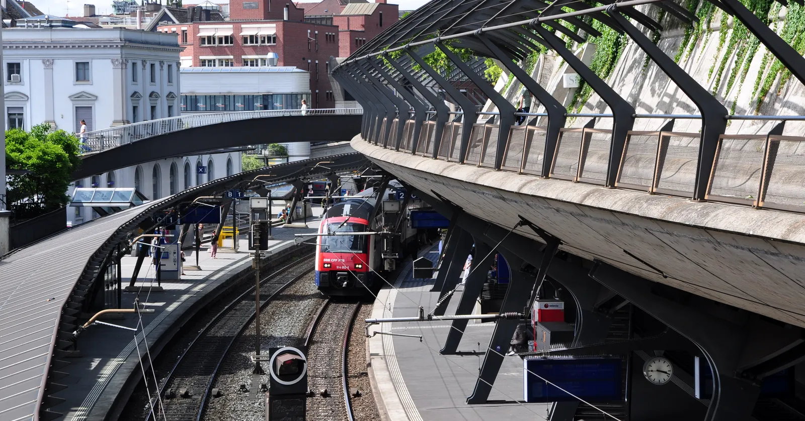 Zürich Stadelhofen station seen from above, with a red train on curved track between platforms and a long ribbed concrete canopy sweeping along the hillside side of the cutting