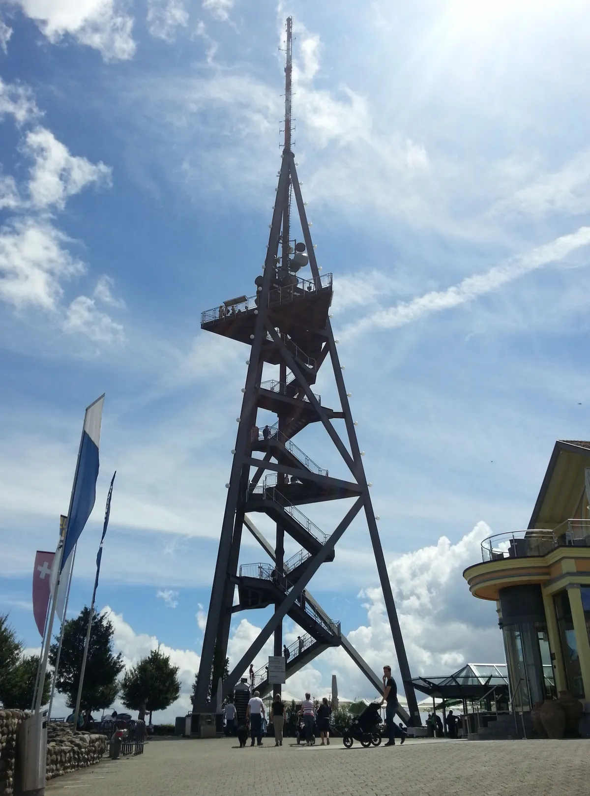 The steel lattice lookout tower on the Uetliberg summit, a tapering four-legged frame with switchback staircases and a railed viewing platform near the top, with flags and visitors on the plateau below it