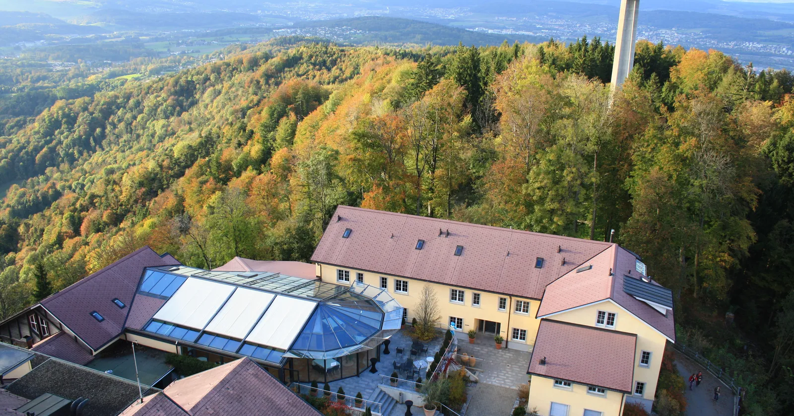 The Uetliberg summit plateau seen from above, with the Uto Kulm hotel and its glass-roofed terrace among pitched roofs, the slim white television mast behind, and wooded ridges and the plain stretching away in autumn colour
