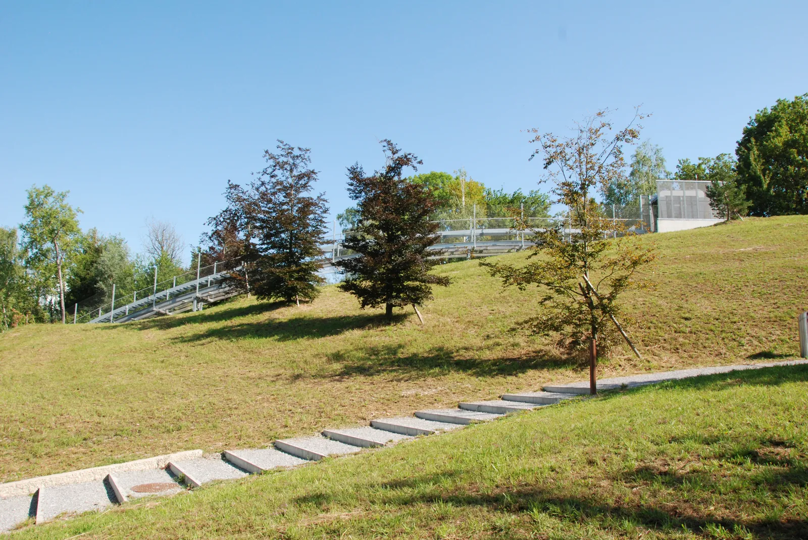 The grassed slope of Circle Park beside Zurich Airport, with young trees, a stepped concrete path climbing the hill and a railed walkway running along the crest against a blue sky