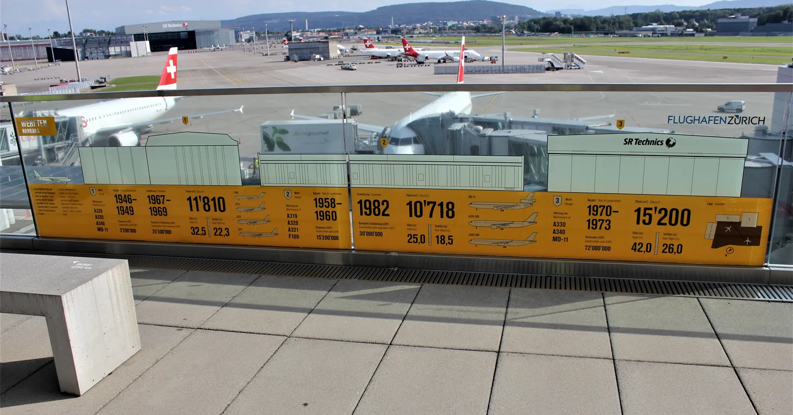 Observation Deck B at Zurich Airport, a paved open terrace with a long glass information panel along its railing and parked airliners, service vehicles and the taxiways of the apron spread out behind it