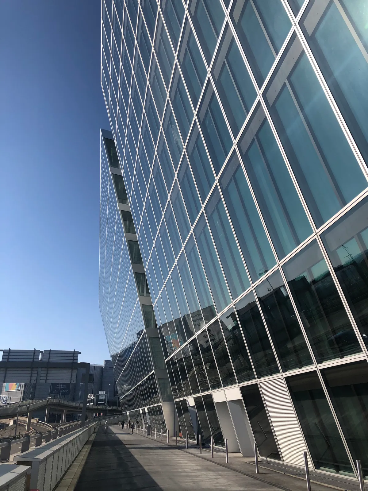 The glass facade of The Circle at Zurich Airport rising above a raised walkway, its angled window grid running back toward the airport terminal buildings in the distance under a clear sky